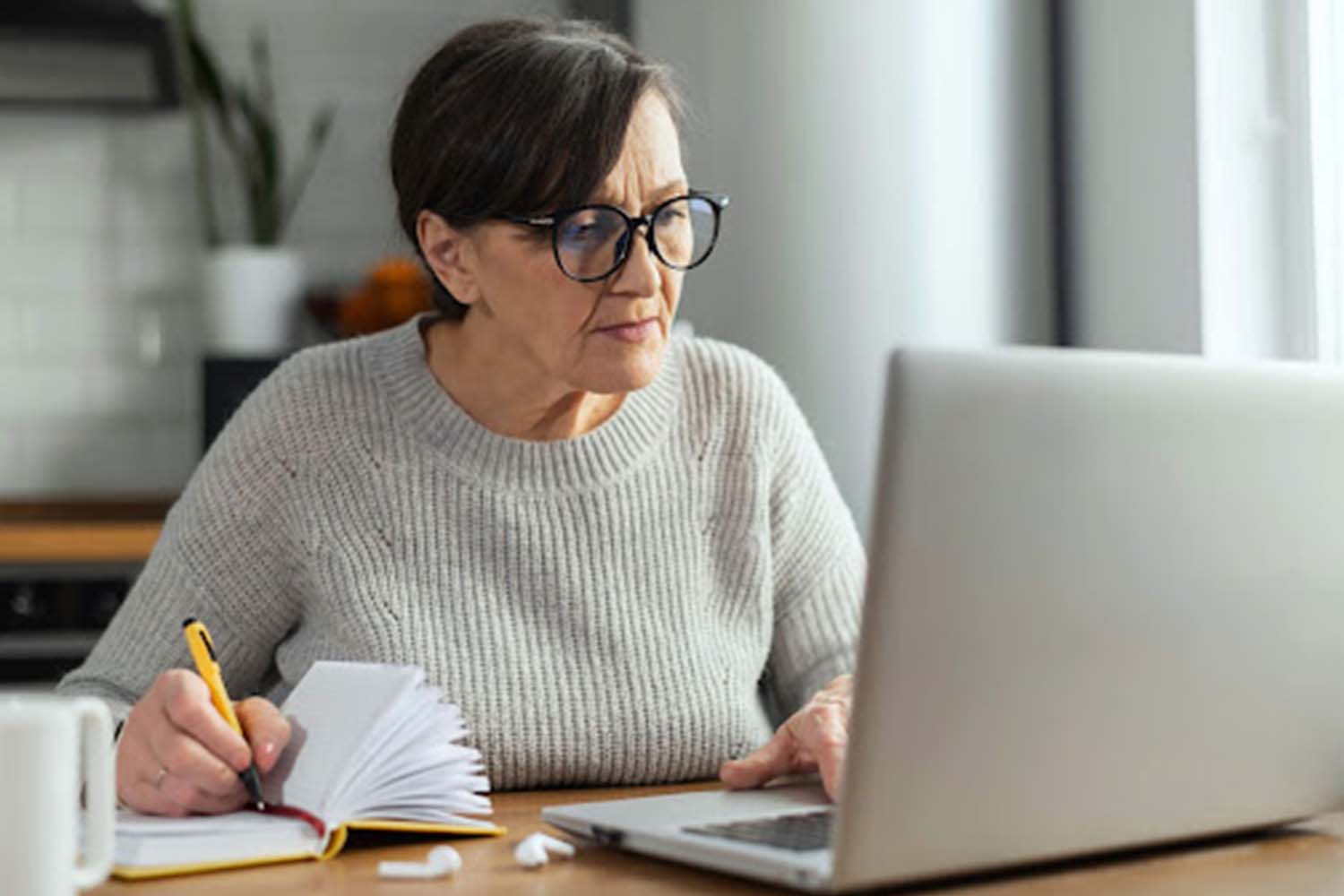 a mature woman transferring notes from a laptop into a notebook